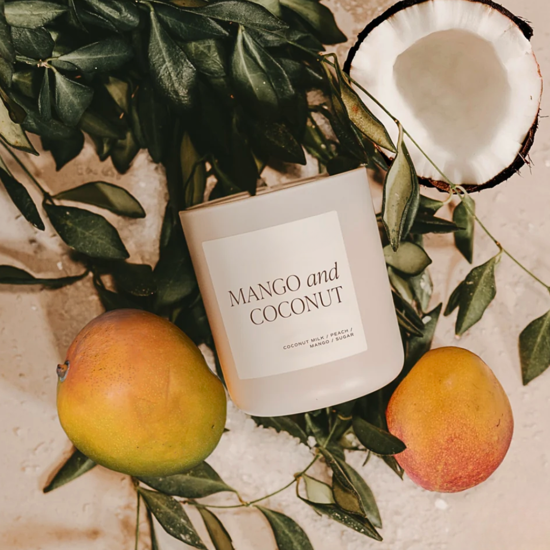 Candle labeled 'Mango and Coconut' surrounded by mangoes and a coconut on a leafy background