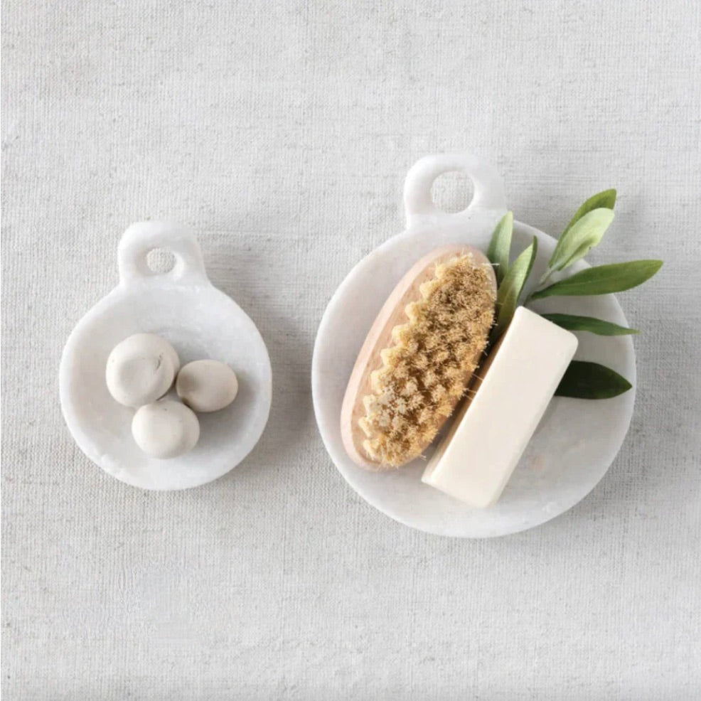 A white marble dish with a round handle, placed on a neutral background, containing what appears to be a round object and a block of marble soap.
