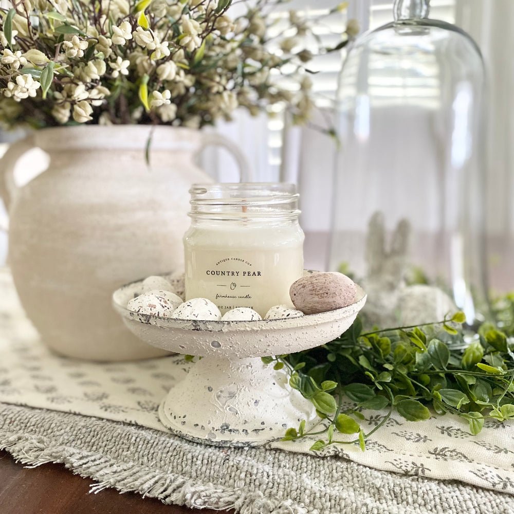A white mason jar Country Pear scented candle with a galvanized metal lid, placed on a decorative white stand, surrounded by a green plant and other home décor elements.