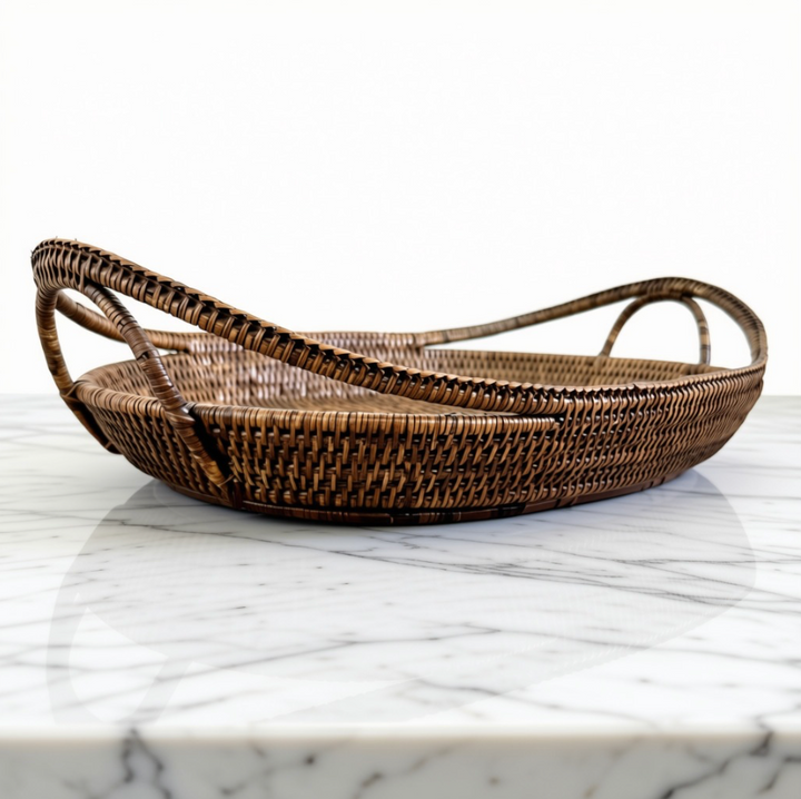An oval woven rattan tray with integrated loop handles, resting on a white marble surface against a white background.
