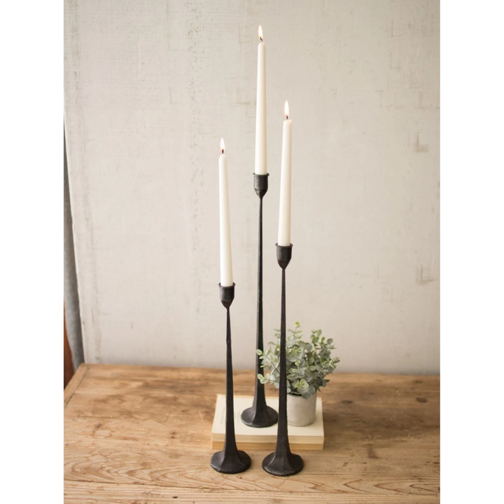 Three black cast iron taper candle holders with white taper candles, styled on a wooden tabletop with a small potted plant against a textured light gray wall.