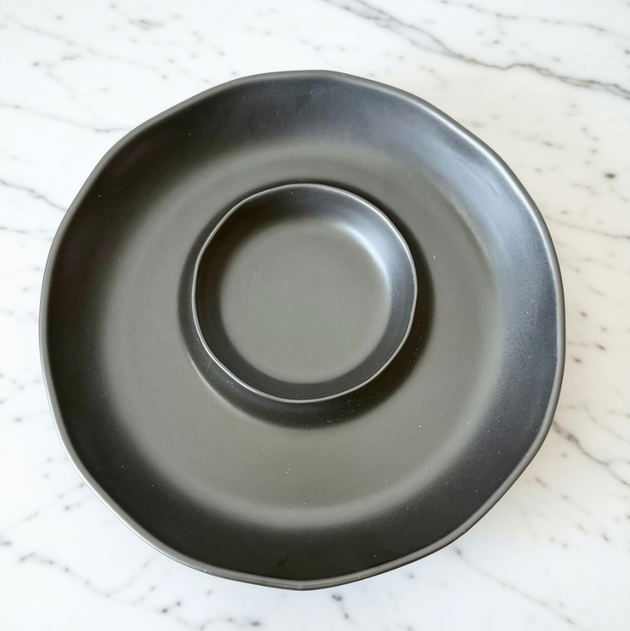 A matte black ceramic chip & dip serving platter with a scalloped rim, photographed from directly above on a white marble surface with gray veining.