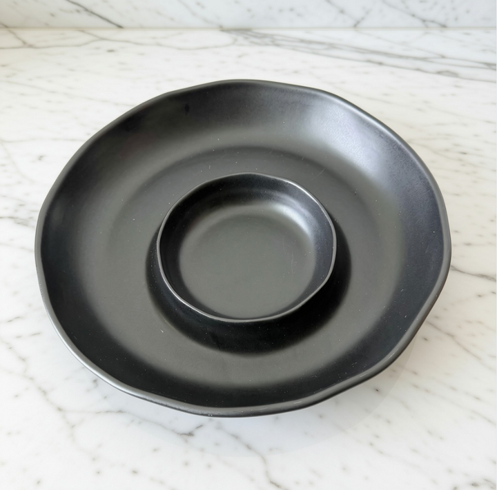 A matte black ceramic dipping platter with a scalloped rim and a central well, shown on a white marble surface.