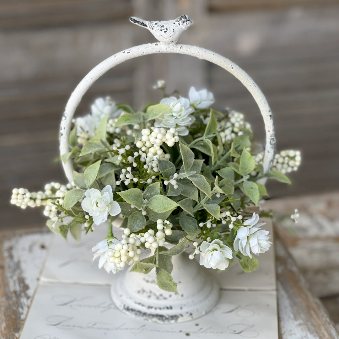 Decorative white metal basket with artificial flowers and a bird ornament on a wooden surface.