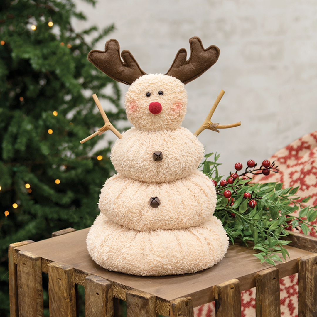 Decorative snowman with reindeer antlers on a wooden crate with a Christmas tree in the background.