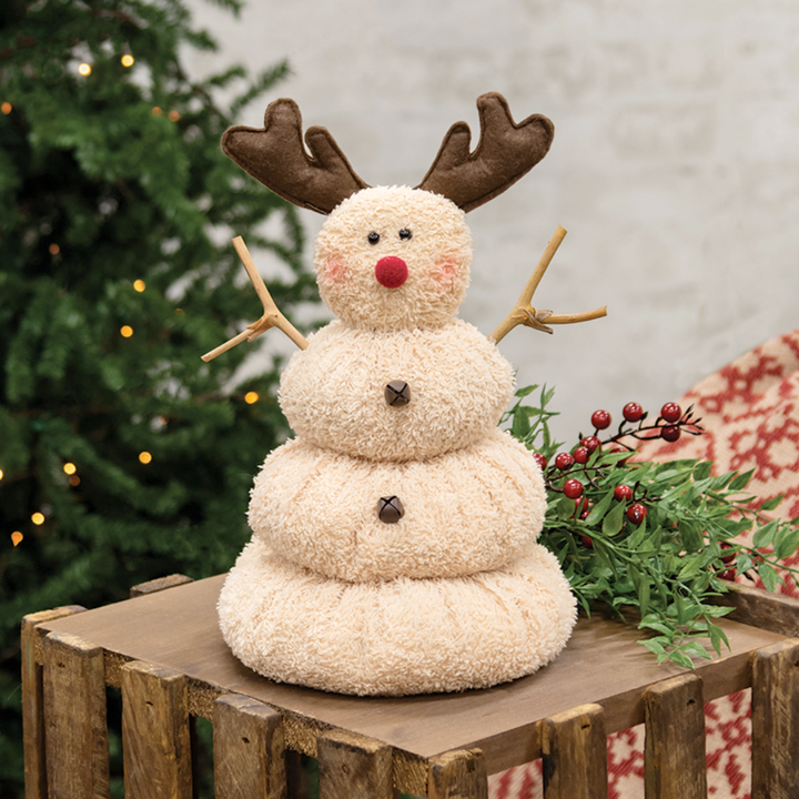 Decorative snowman with reindeer antlers on a wooden crate with a Christmas tree in the background.