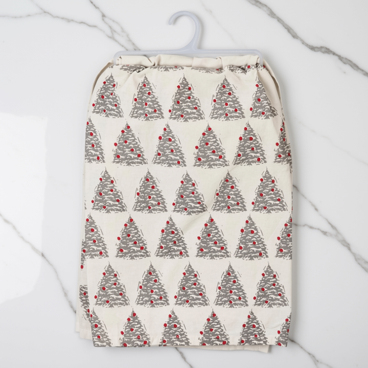 A kitchen tea towel with a repeating Christmas tree pattern in gray and red, hanging on a white plastic hanger against a white marble background.