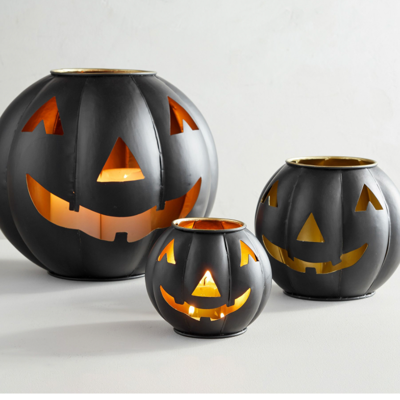 Three black pumpkin-shaped candle holders with carved jack-o'-lantern faces, arranged in a size progression on a light gray background. Each holder is lit from within, casting a warm orange glow.