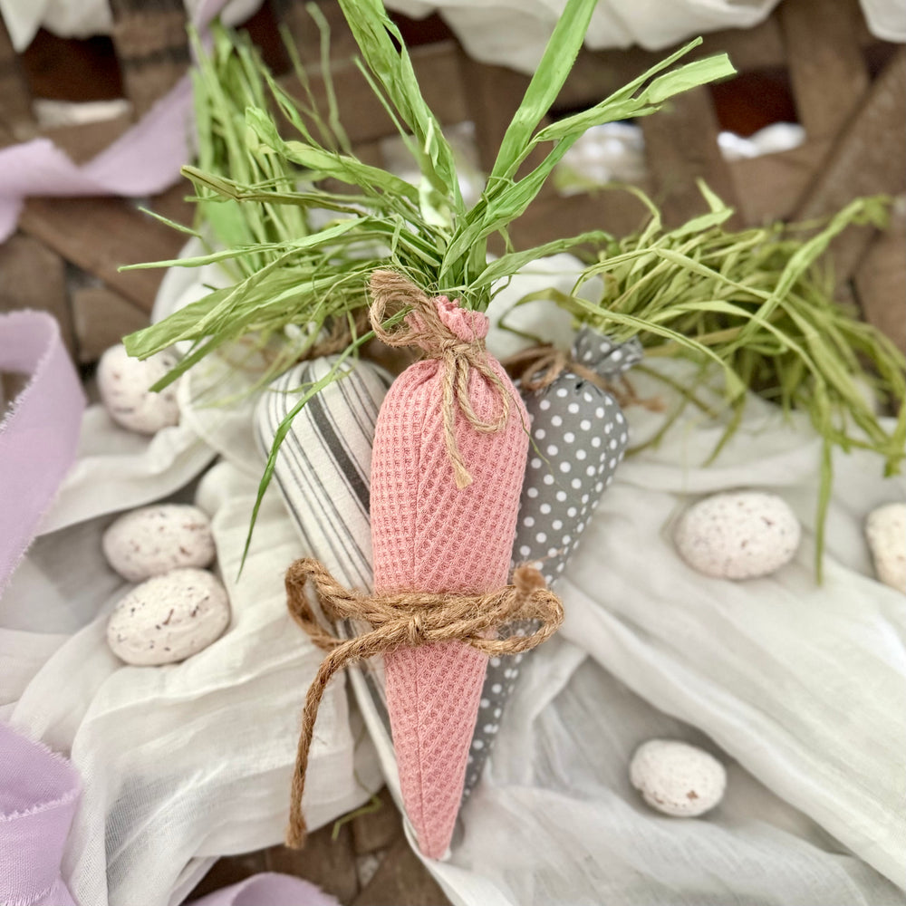 Decorative carrots made of pink and gray fabrics with green raffia leaves on a textured surface.
