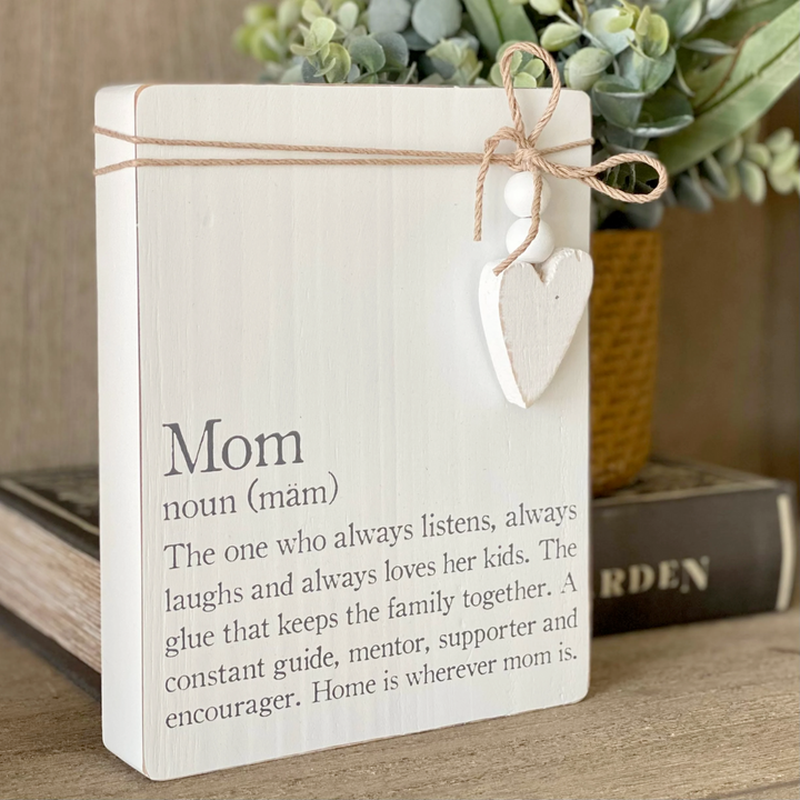 A wooden plaque with a definition of 'Mom' written on it, adorned with twine, wooden beads, and a heart charm, placed on a wooden surface with a plant in the background.