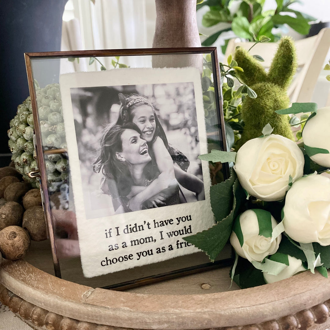 A brass finished metal picture frame with pressed glass panes displaying a photo and a parchment paper matting with a sentimental message.
