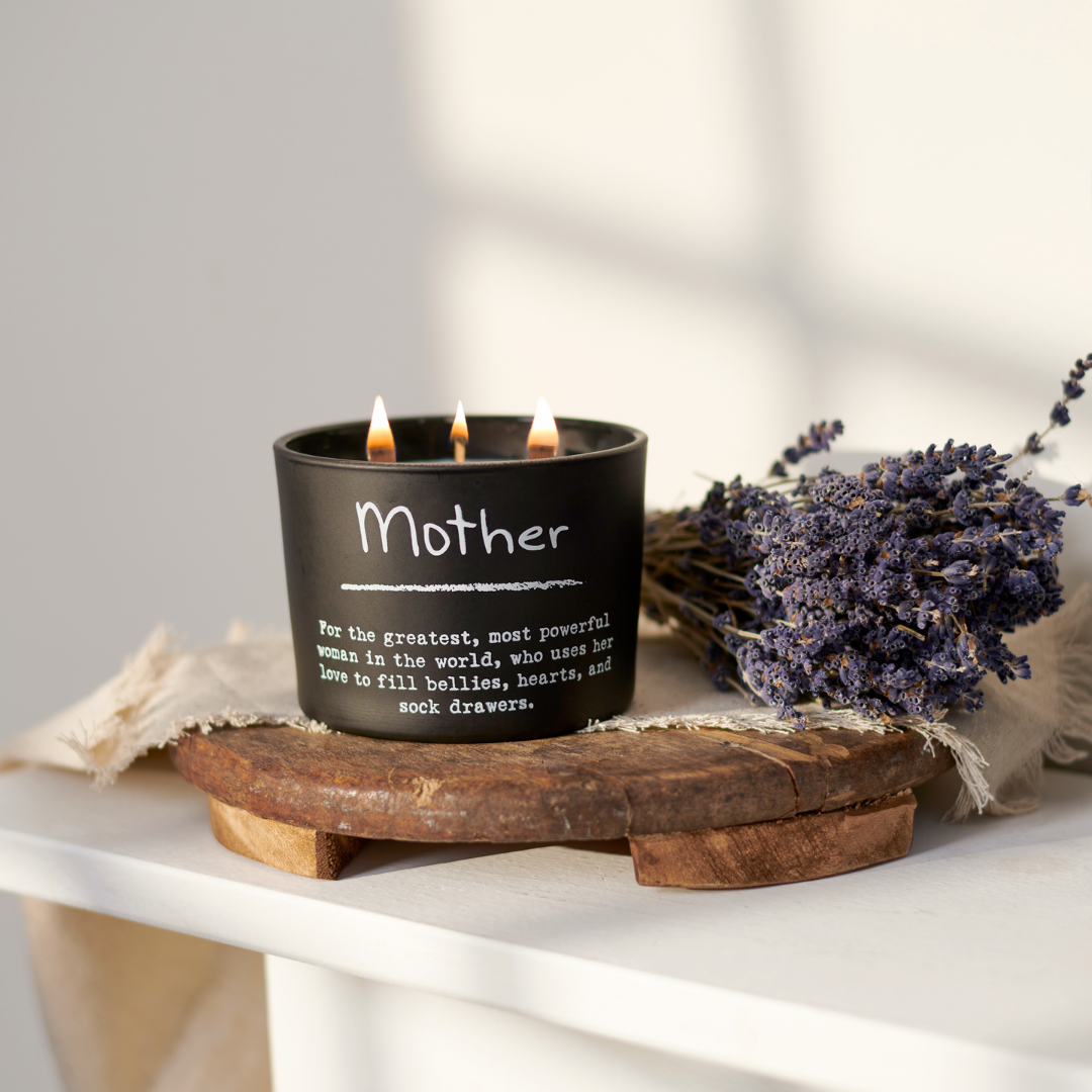 A matte-black glass jar with a triple-wick lavender scented candle, placed on a wooden surface with a protective plastic wax cover, surrounded by dried lavender and a burlap napkin with the word 'Mother' printed on it.