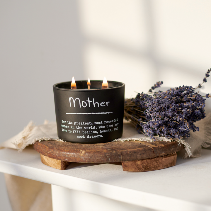 A matte-black glass jar with a triple-wick lavender scented candle, placed on a wooden surface with a protective plastic wax cover, surrounded by dried lavender and a burlap napkin with the word 'Mother' printed on it.