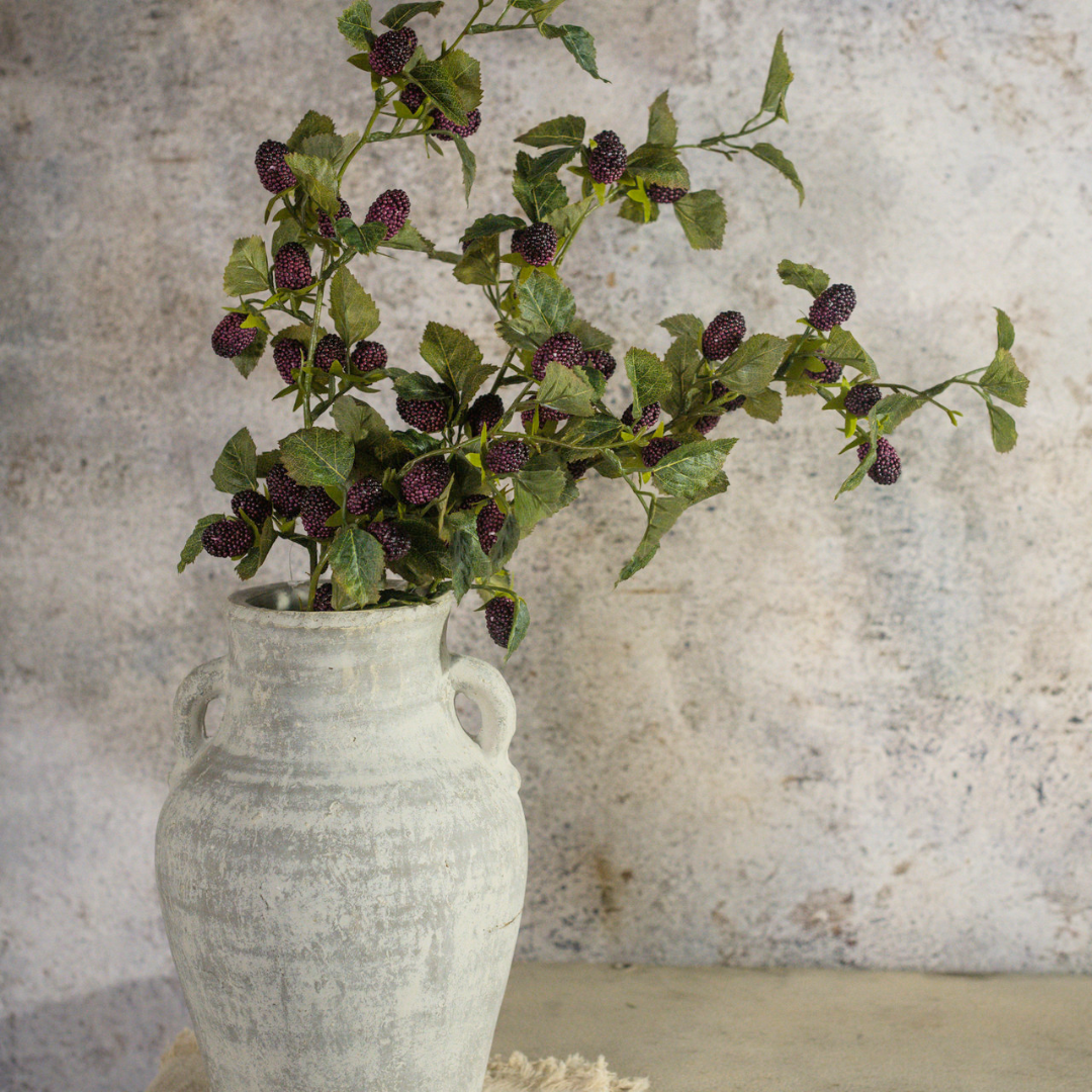 Artificial mulberry sprays with purple berries in a white vase against a textured wall.