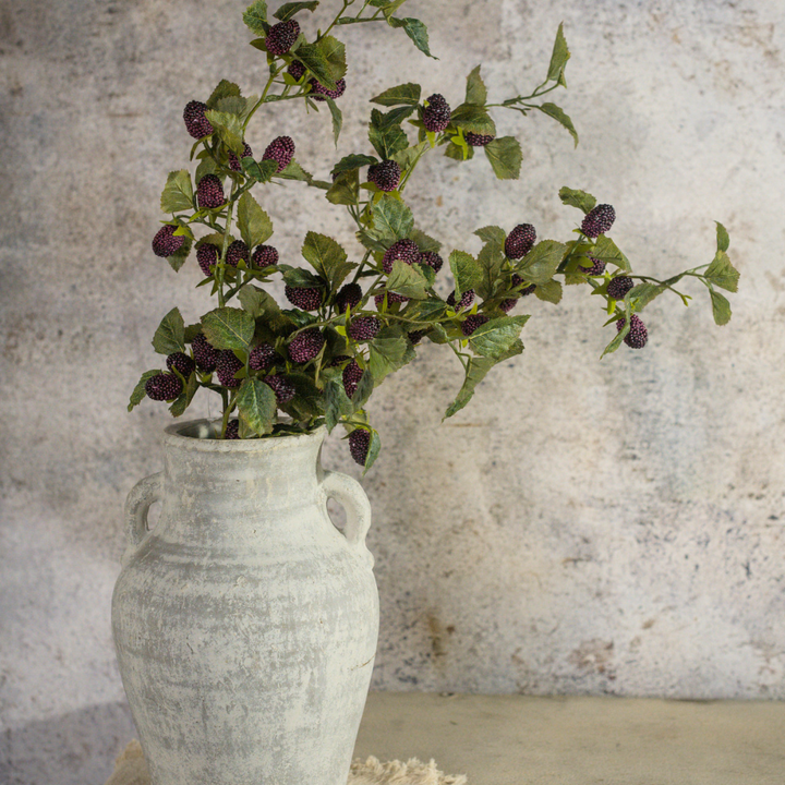 Artificial mulberry sprays with purple berries in a white vase against a textured wall.