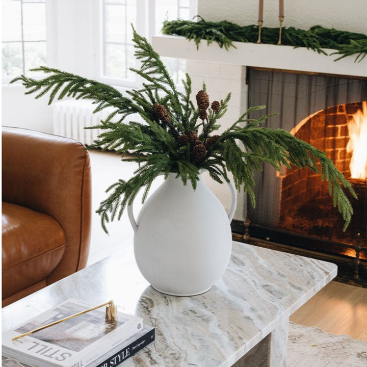 White vase with greenery on a marble table in a living room with a fireplace.