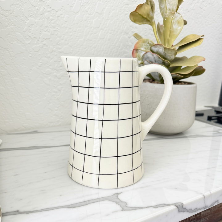 An off-white stoneware pitcher with a black grid pattern, sitting on a white marble countertop with a potted succulent in the background.