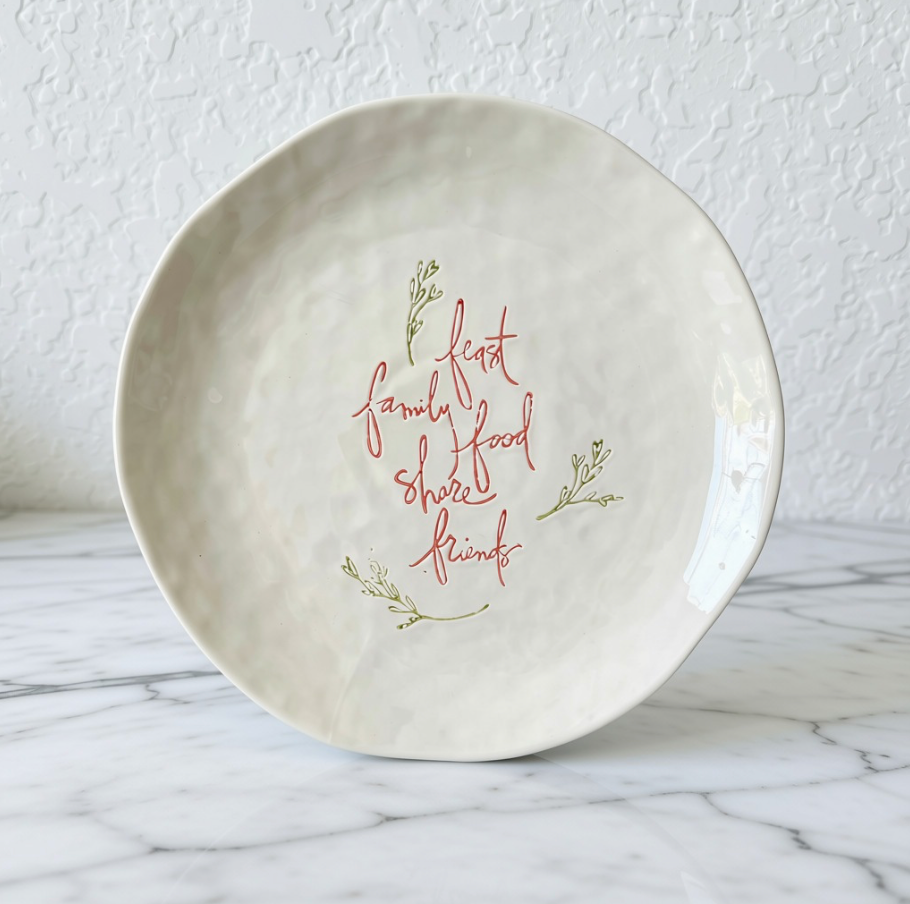 An off-white stoneware serving platter with hand-painted red and green script reading 'feast family food share friends', displayed on a white marble surface against a textured white wall.