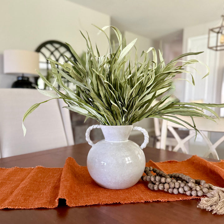 White vase with greenery on a table with an orange runner