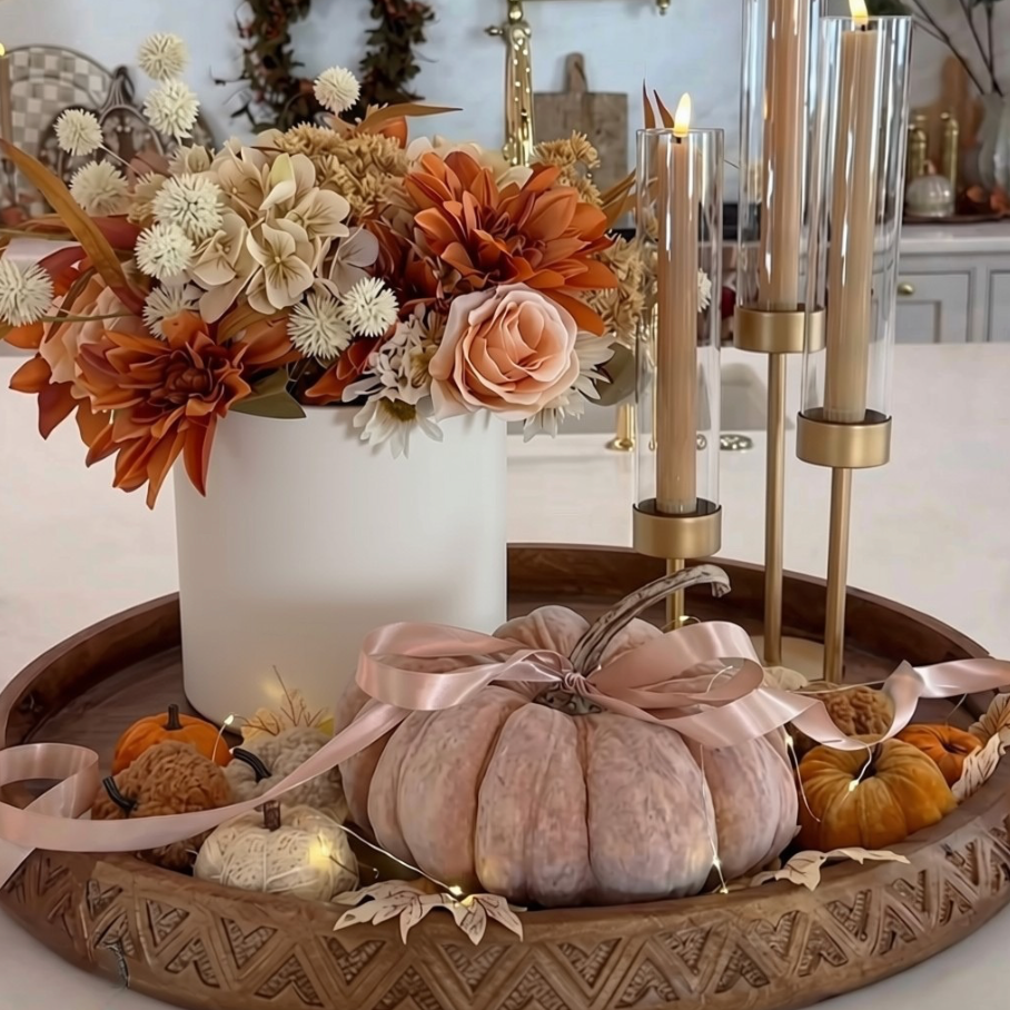A styled autumn centerpiece on a carved wooden tray, featuring a white vase filled with dried orange, beige, and cream flowers, a large pale pink decorative pumpkin with a satin ribbon, small orange pumpkins, fall leaves, and lit gold candlesticks in glass holders.