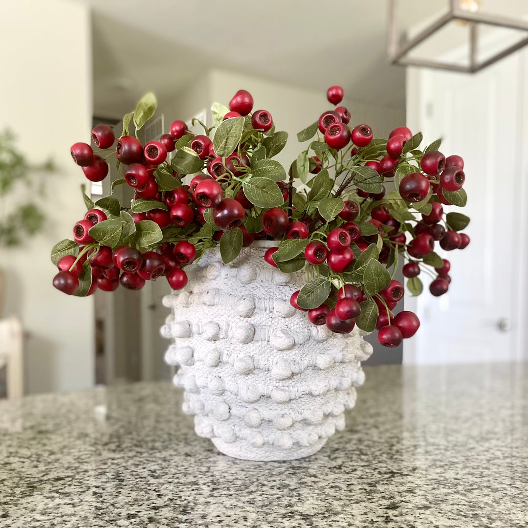 A faux red pod stem with green leaves arranged in a white vase, placed on a counter with a striped napkin beside it.