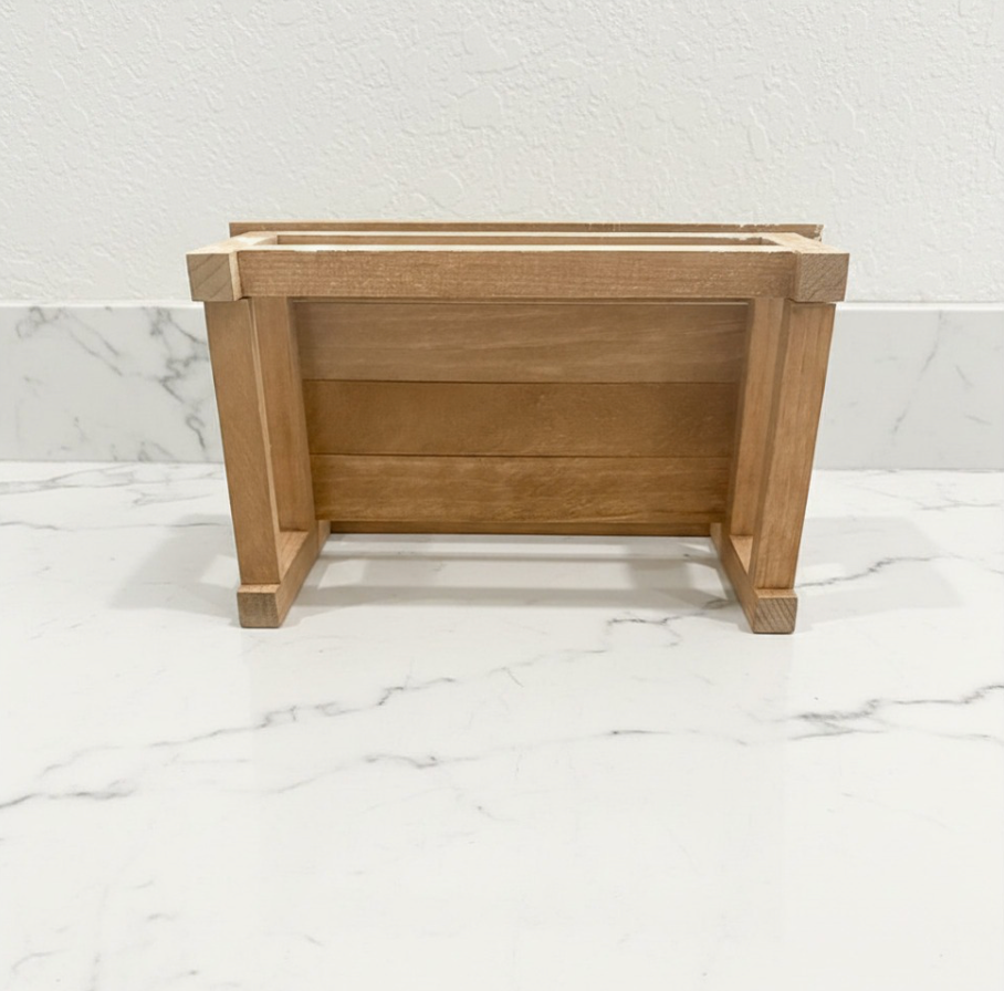 A small wooden planter with a natural oak finish, photographed from the front on a white marble surface against a white textured wall.