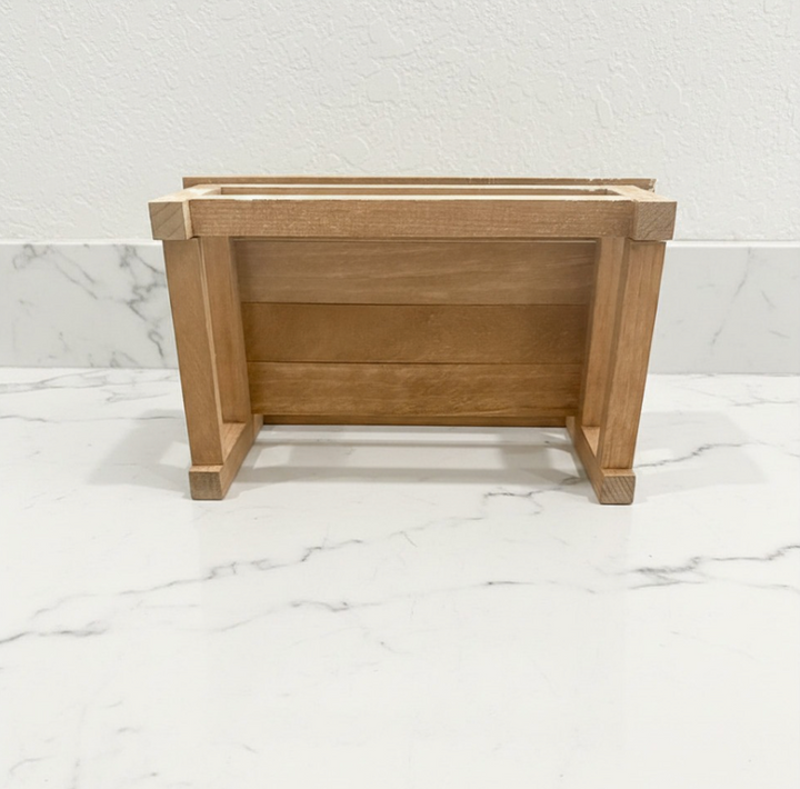 A small wooden planter with a natural oak finish, photographed from the front on a white marble surface against a white textured wall.