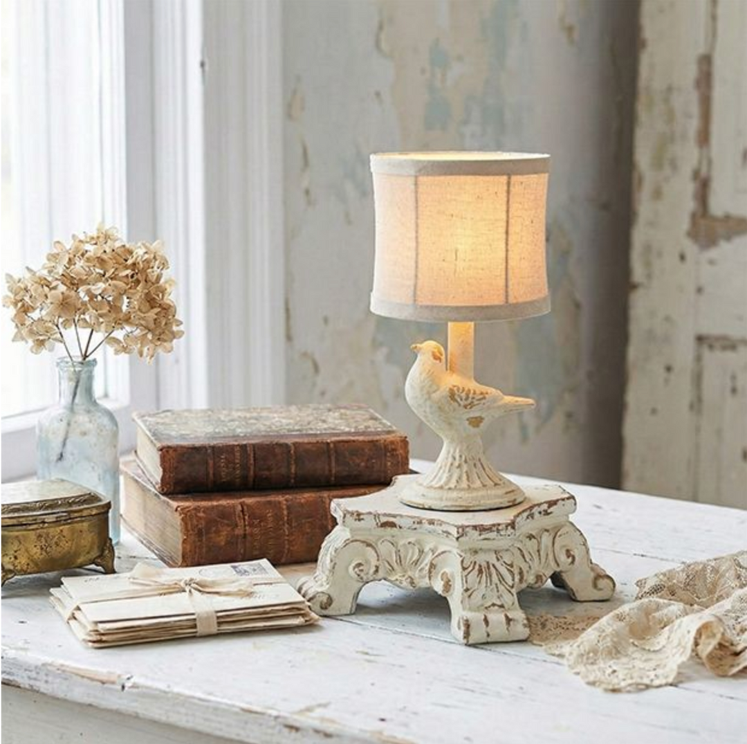 A decorative table lamp with a bird-shaped sculpted base and a drum-style fabric shade, shown lit on a distressed white pedestal riser in a softly styled interior setting with books, a vase of dried flowers, and lace.