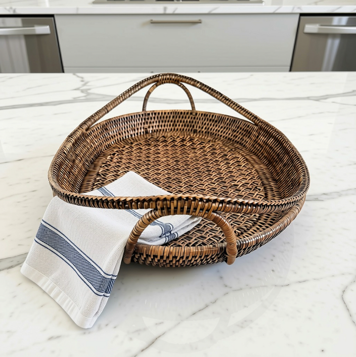 An oval woven rattan serving tray with two loop handles sits on a white marble kitchen countertop, with a folded white kitchen towel draped over one handle.
