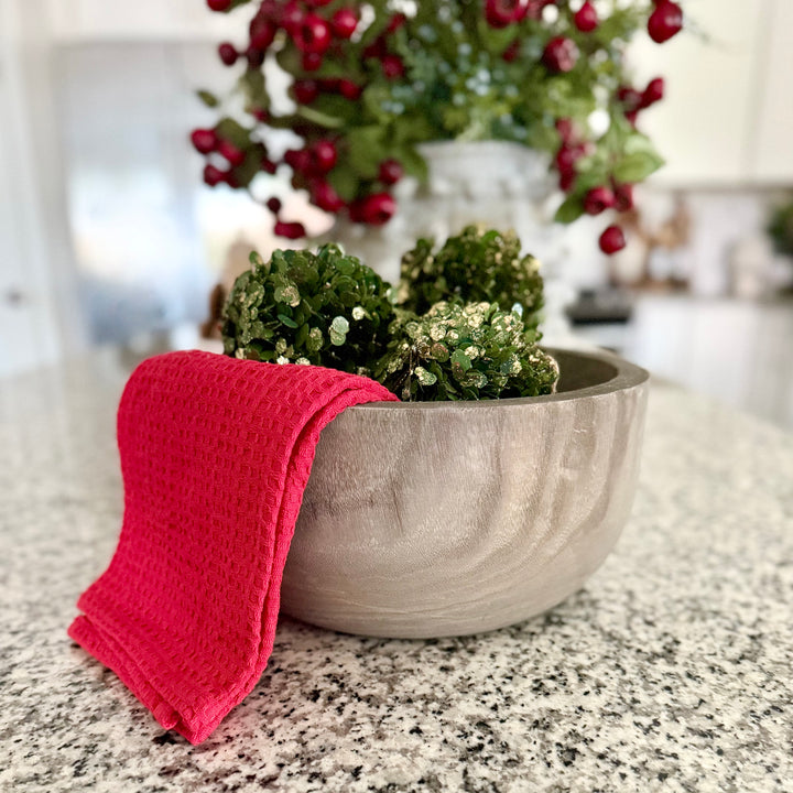 Decorative wood bowl with green spherical ornaments and a red kitchen towel on a countertop, with a blurred festive background.