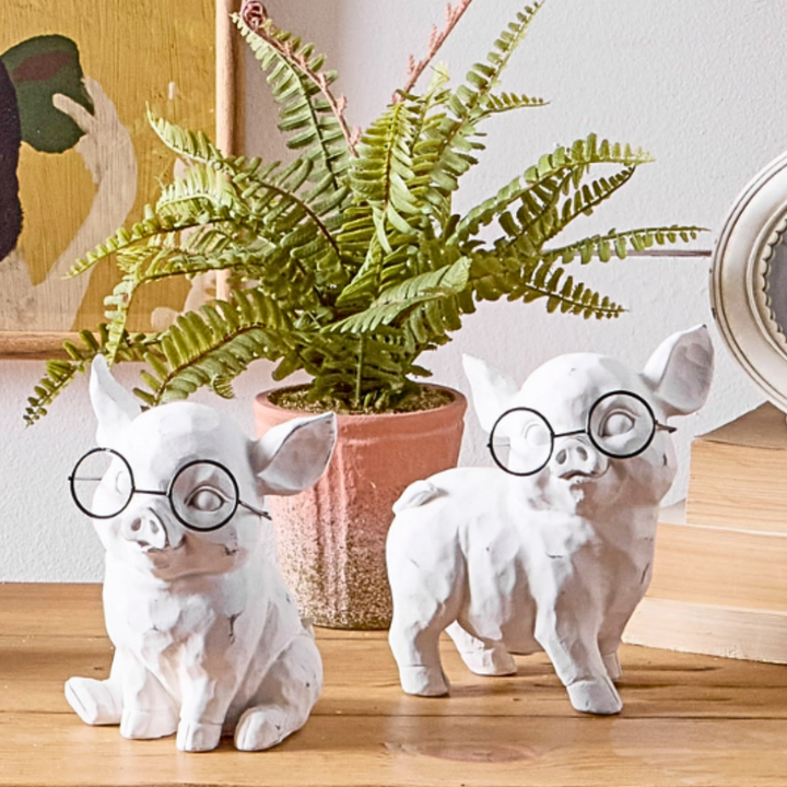 Two white ceramic pig figurines with eyeglasses on a wooden surface with a plant in the background.