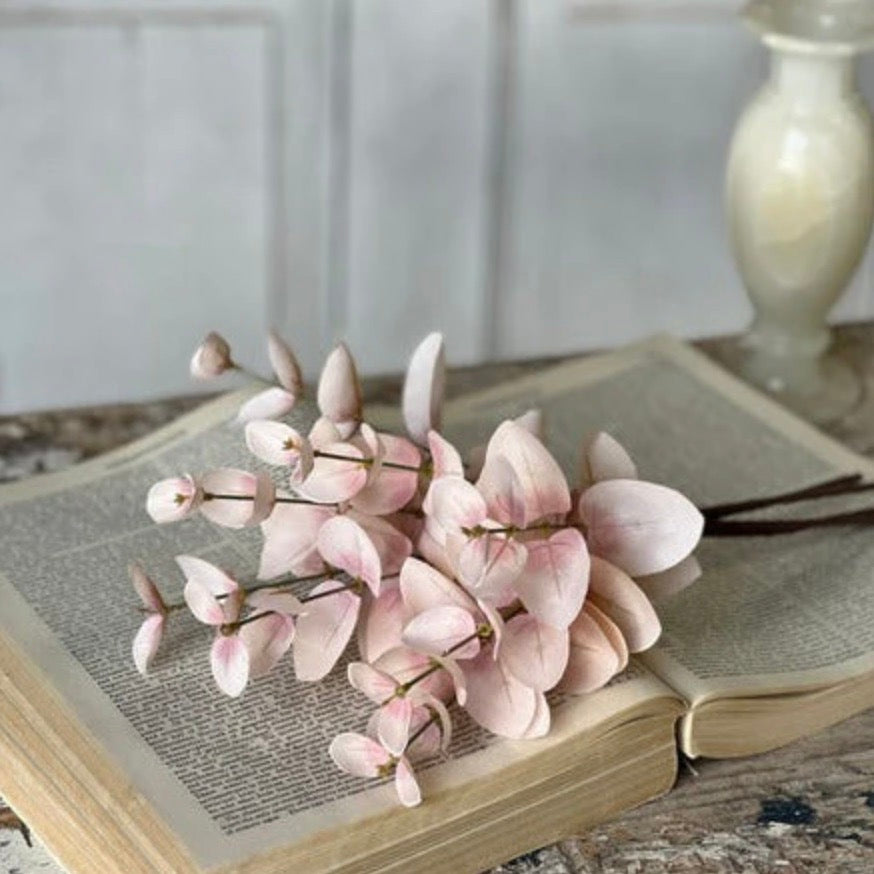 A set of three artificial pink eucalyptus sprays placed on an open book, with a decorative vase in the background.