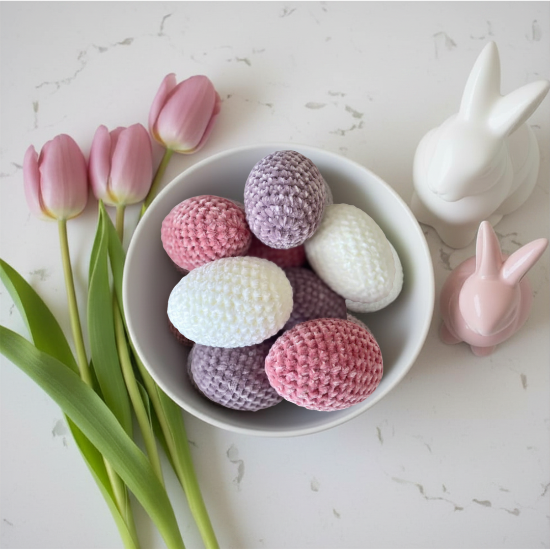 White bowl with pink, purple, and white crocheted eggs, tulips, and rabbit figurines on a light surface.