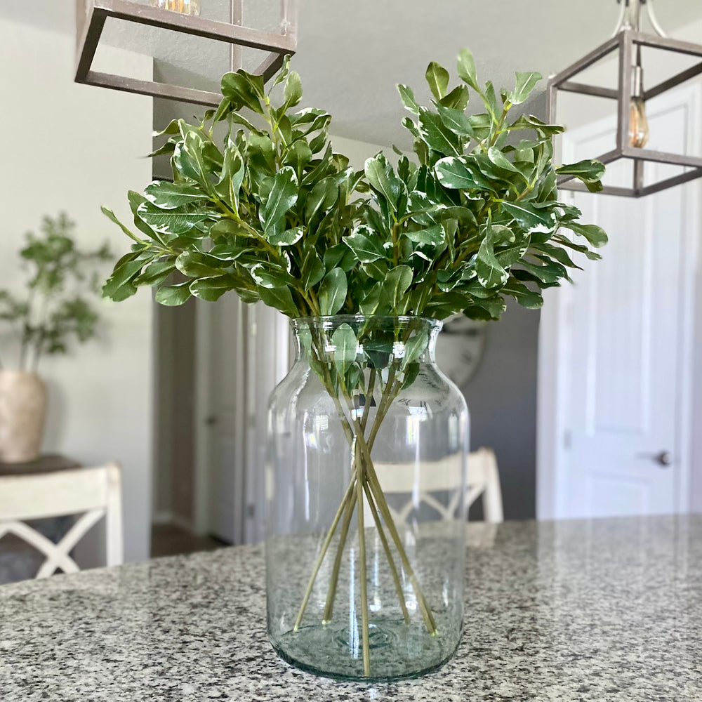 A lifelike greenery stem with deep green leaves in a clear glass vase, placed on a kitchen countertop.