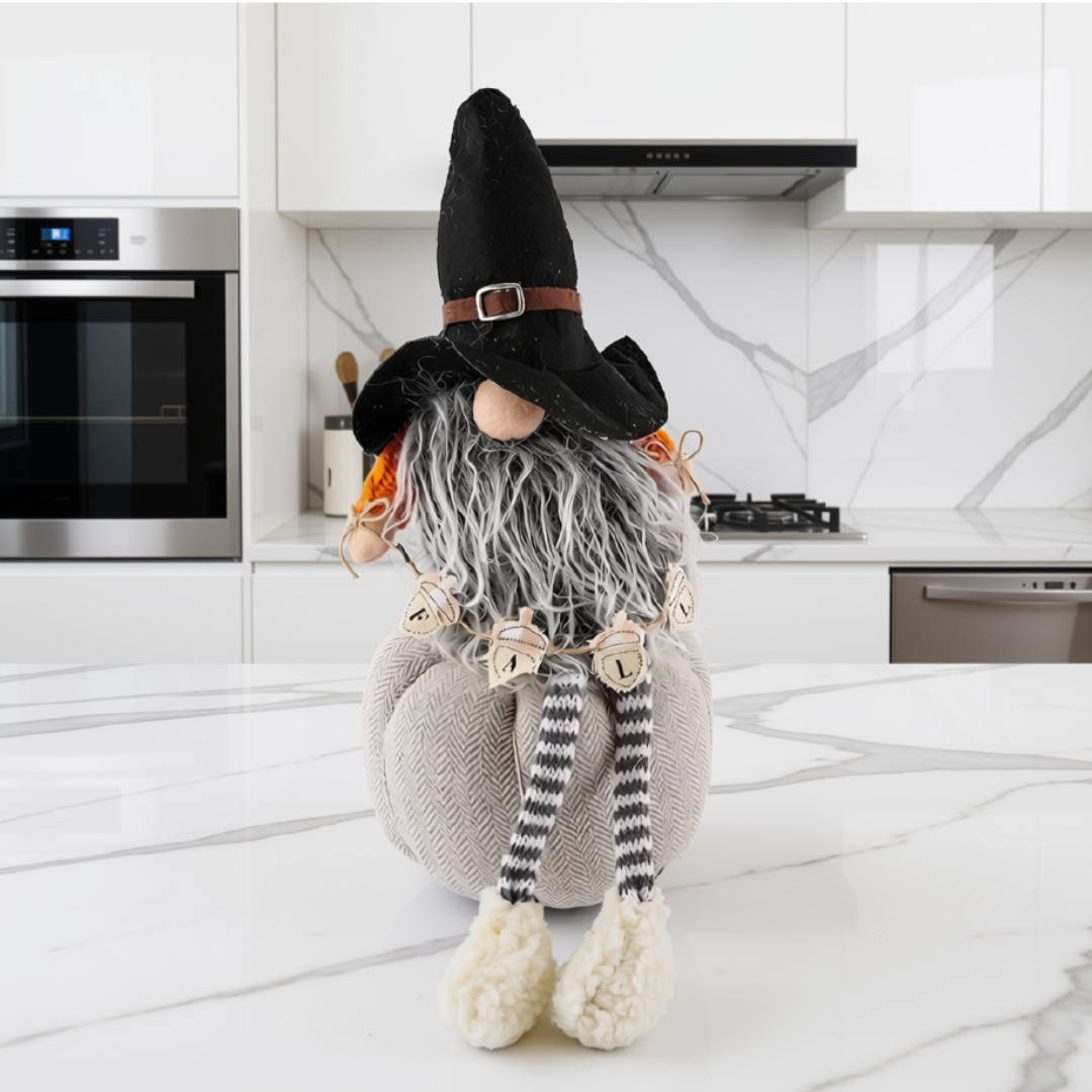 A decorative pumpkin gnome doll with a black hat, gray beard, striped legs, and cream shoes, posed on a white marble kitchen countertop.