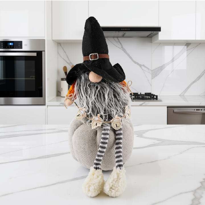 A decorative pumpkin gnome doll with a black hat, gray beard, striped legs, and cream shoes, posed on a white marble kitchen countertop.
