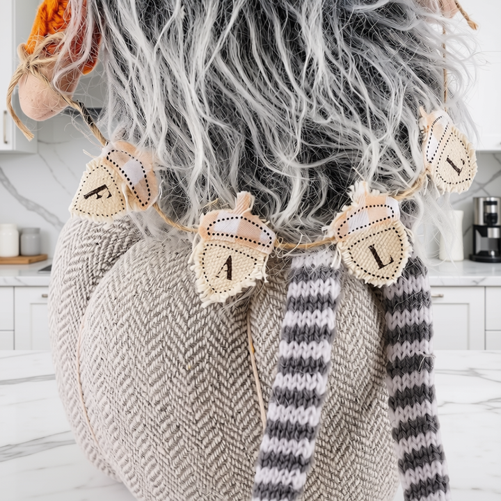 A close-up of a person wearing a chunky beige herringbone knit sweater with a gray and white striped knit scarf, styled with a fluffy gray wig and a decorative autumn leaf garland.