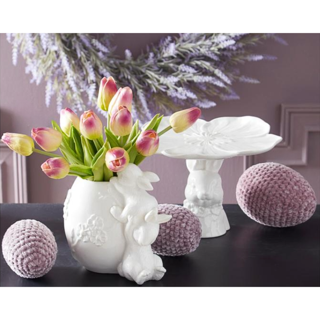 Decorative scene with a white vase shaped like a rabbit with pink tulips, purple textured Easter eggs, and a white table.