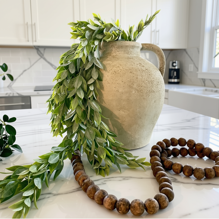 A large beige ceramic vase filled with cascading green leafy branches, styled on a white marble kitchen counter with a wooden bead necklace in the foreground.