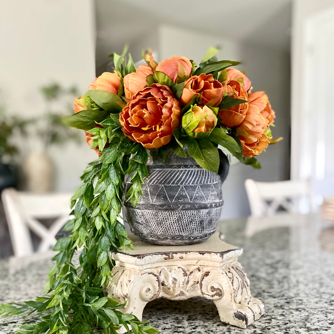 A bundle of artificial peony blooms in an unfurled condition, tied with raffia, displayed in a gray vase with a white ornamental base on a kitchen counter.