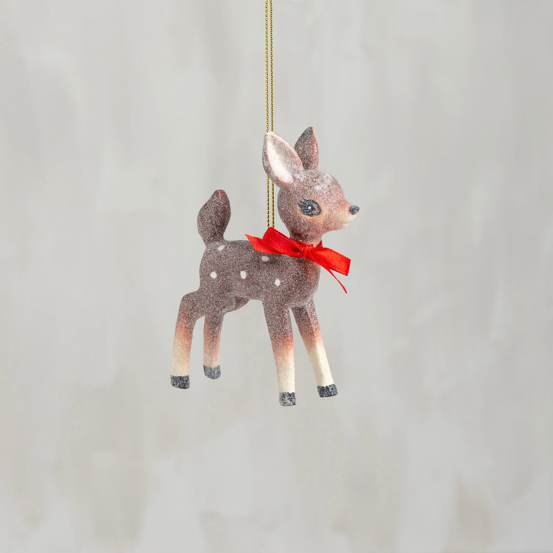 A vintage-style deer ornament with a red bow, hanging from a chain against a neutral background.