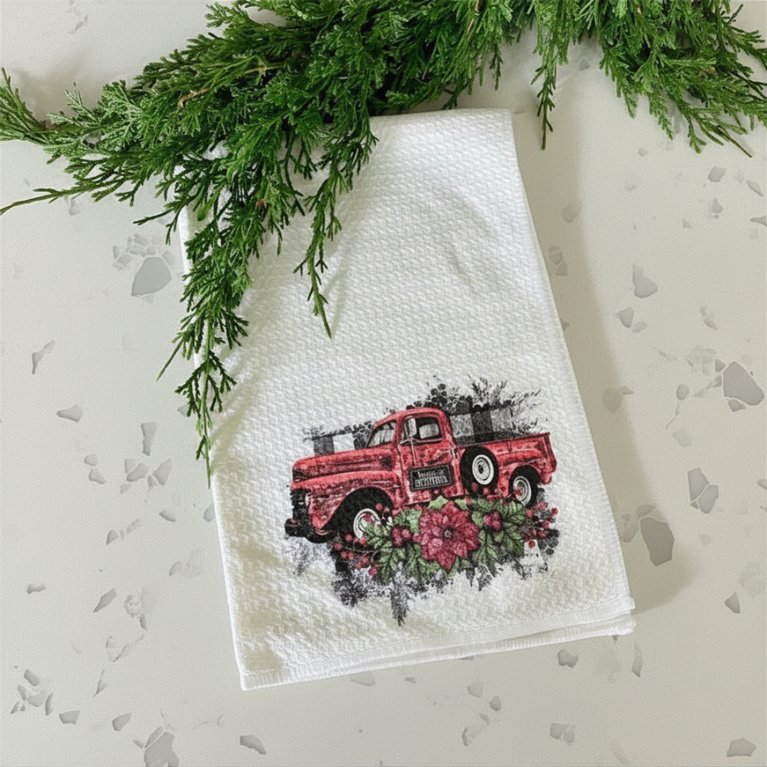 White towel with a red truck and floral design, surrounded by greenery on a light background