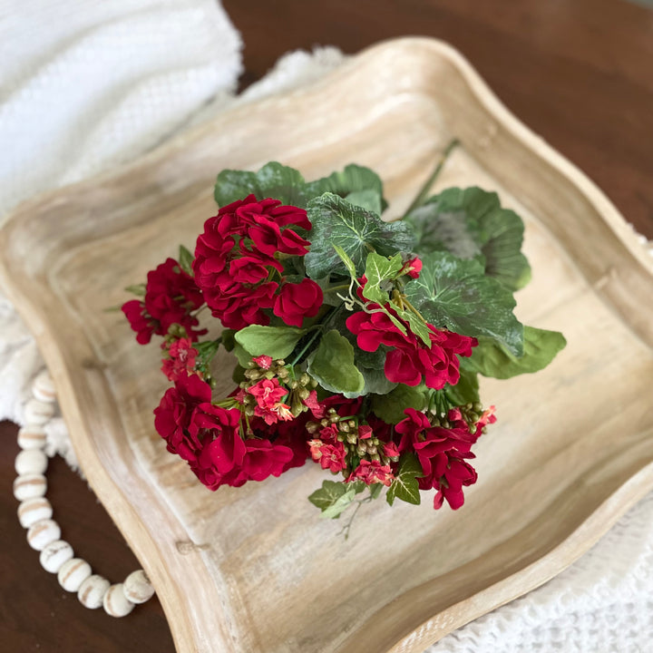 A decorative woven pitcher with a lifelike arrangement of red geranium and kalanchoe bush in it, placed on a wooden surface.