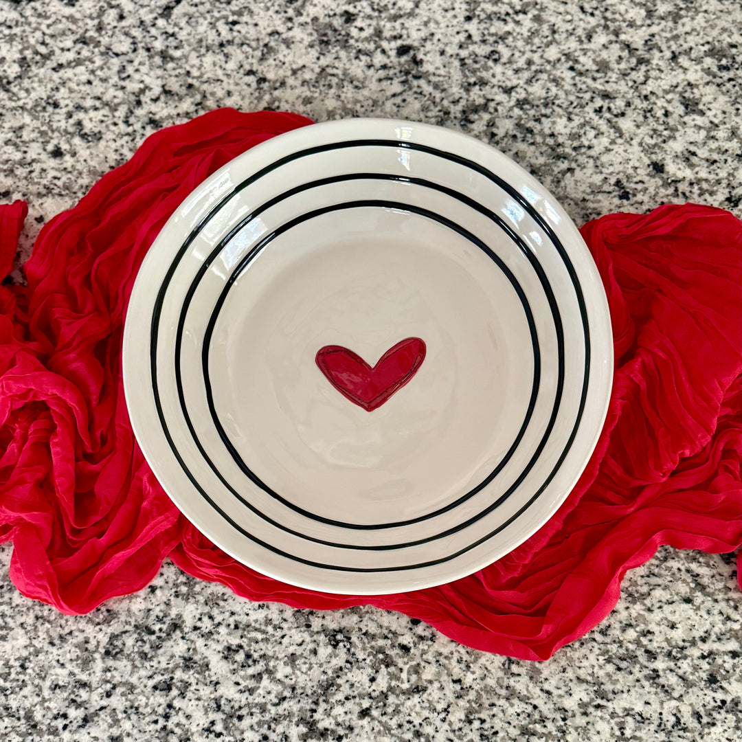 White stoneware serving bowl with black stripes and a red heart on a red cloth over a gray granite surface