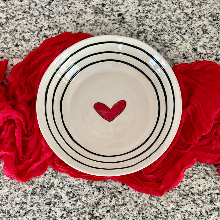 White stoneware serving bowl with black stripes and a red heart on a red cloth over a gray granite surface