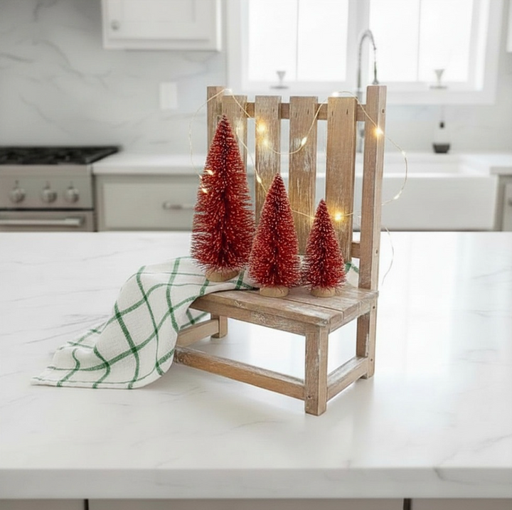 A rustic wooden chair-style decorative stand holding three red bottle tree ornaments with warm fairy lights draped around them, set on a white kitchen countertop with a green-and-white checkered cloth beside it.