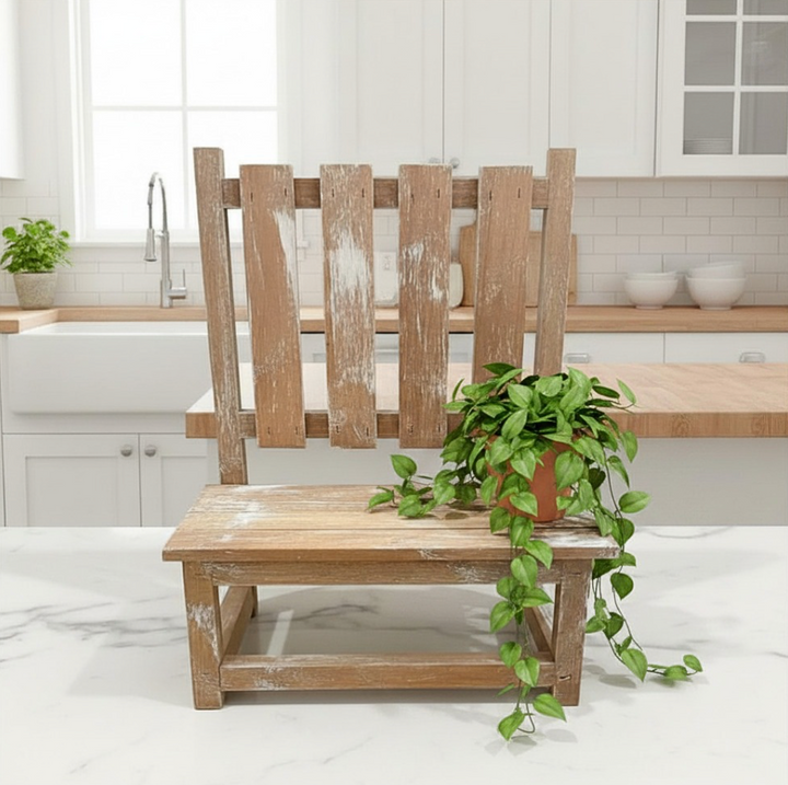 A rustic wooden bench with a distressed finish stands on a white marble countertop in a bright, modern kitchen. A green trailing plant in a terracotta pot is placed on the chair's seat.