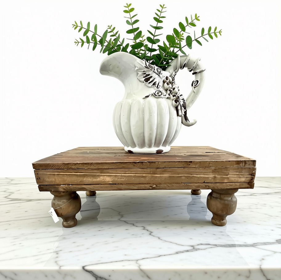 A white ribbed ceramic pitcher vase with a decorative silver-toned floral accent on the handle, filled with green foliage, displayed on a Footed Serving Stand crafted from rustic planked pine wood and features distinctive hand-turned pedestal feet over a white marble surface against a white background.