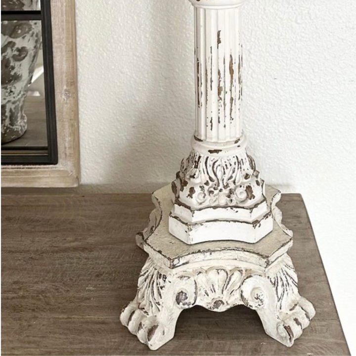 A distressed white decorative column base with ornate carved details and claw-like feet, shown on a wooden surface against a white wall.