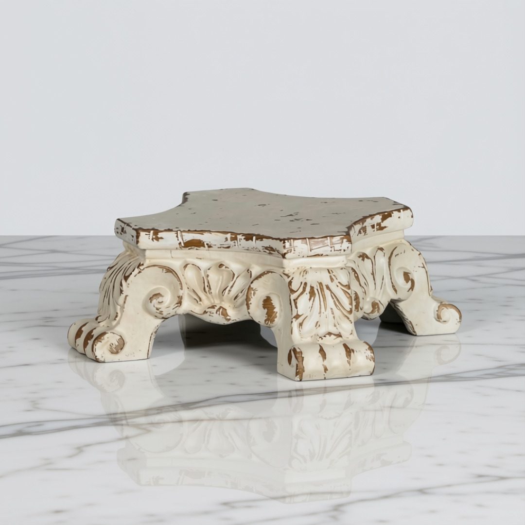 An ornate, distressed white wooden stand or miniature pedestal with carved scrollwork and clawed feet, resting on a white marble surface with a light gray background.